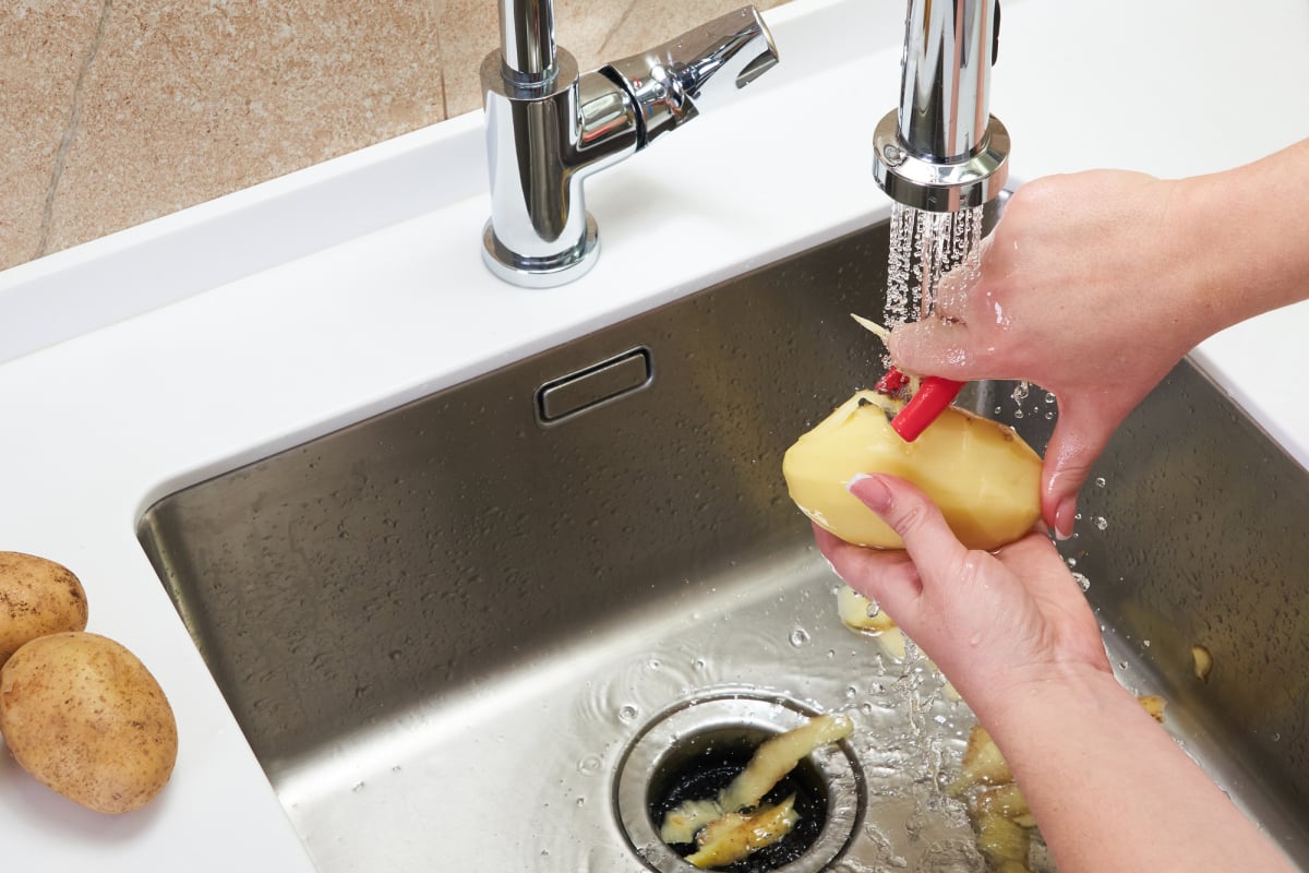 Cropped view of female hands peeling potato over Food waste disposer machine in sink in modern kitchen